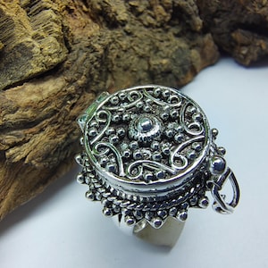 Op de afbeelding: Een zilverkleurige ring met een ronde medaillon-bovenzijde, voorzien van een gedetailleerd reliëfpatroon van krullende lijnen en kleine verhoogde bolletjes. Het medaillon heeft een scharnier aan de zijkant en een decoratieve geschulpte rand.