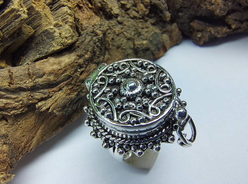 May include: A silver-toned ring featuring a circular locket top with an intricate embossed pattern of swirling lines and small raised spheres. The locket has a hinged side and a decorative scalloped edge with a textured finish around the base.