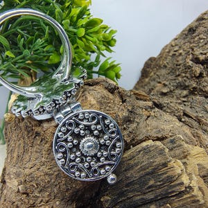 May include: A silver-toned metal ring featuring a circular locket attachment. The locket face displays an intricate embossed pattern of swirling lines and small raised dots, with a central circular stud and a small hinge at the top.