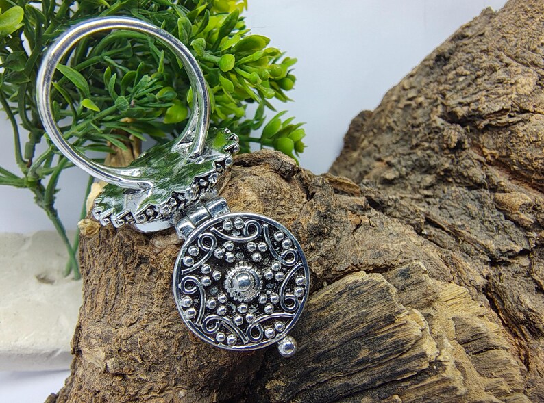 May include: A silver-toned metal ring featuring a circular locket attachment. The locket face displays an intricate embossed pattern of swirling lines and small raised dots, with a central circular stud and a small hinge at the top.