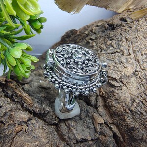 May include: A silver-toned ring featuring a large, circular, hinged locket top with intricate embossed scrollwork and beaded detailing. The metal surface has a polished finish with darkened recessed areas to highlight the ornate, textured pattern.