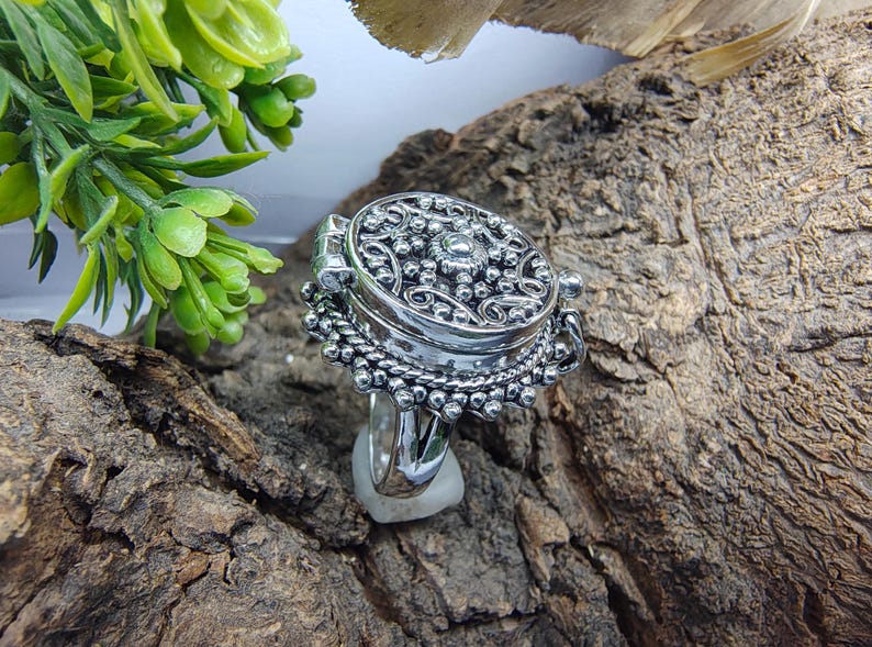 May include: A silver-toned ring featuring a large, circular, hinged locket top with intricate embossed scrollwork and beaded detailing. The metal surface has a polished finish with darkened recessed areas to highlight the ornate, textured pattern.
