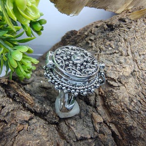 May include: A silver-toned ring featuring a circular locket top with an intricate embossed floral and beaded pattern. The locket is hinged on one side and sits atop a decorative base with a beaded edge, showcasing a polished metallic finish.