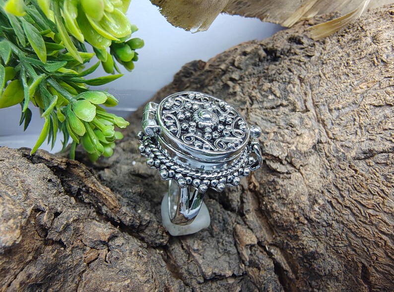 May include: A silver-toned ring featuring a circular locket top with an intricate embossed floral and beaded pattern. The locket is hinged on one side and sits atop a decorative base with a beaded edge, showcasing a polished metallic finish.
