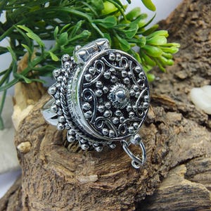 May include: A silver-toned metal ring featuring a large, circular, hinged locket top. The surface of the locket is decorated with intricate embossed scrollwork, small raised spheres, and a central stud, finished with a beaded edge detail.
