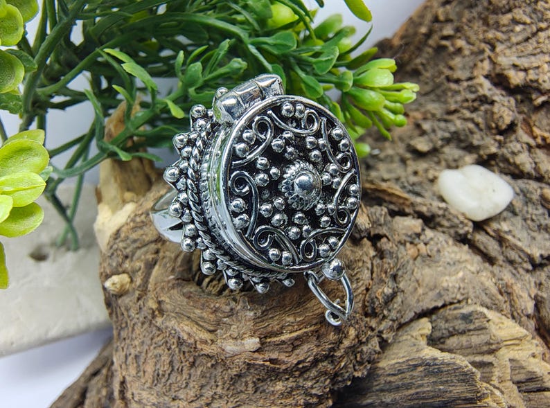 May include: A silver-toned metal ring featuring a large, circular, hinged locket top. The surface of the locket is decorated with intricate embossed scrollwork, small raised spheres, and a central stud, finished with a beaded edge detail.