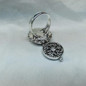 May include: A silver ring featuring a circular setting with a scalloped edge and a dangling, round, filigree-style charm. The charm displays an intricate, perforated metal pattern with a small, central, raised silver bead detail.