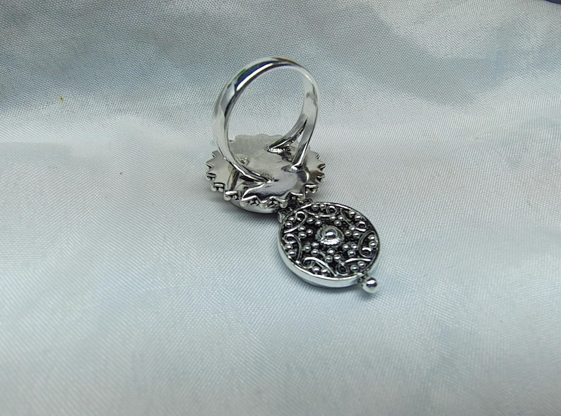 May include: A silver ring featuring a circular setting with a scalloped edge and a dangling, round, filigree-style charm. The charm displays an intricate, perforated metal pattern with a small, central, raised silver bead detail.
