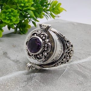Op de afbeelding: Een zilverkleurige metalen medaillonring met een cirkelvormig voorvlak en een centrale ronde paarse edelsteen. Het medaillon heeft een scharnierend deksel met decoratief metaalwerk, inclusief kraaldetails en krulpatronen op de zijkanten.