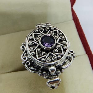 Puede incluir: Un anillo guardapelo de plata con una cara circular que presenta un intrincado patrón en relieve de corazones y pequeñas esferas. Una piedra preciosa redonda y facetada de color púrpura está engastada en el centro, con una bisagra visible.