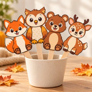 May include: Four cartoon animal cutouts featuring a fox, an owl, a bear, and a deer, each attached to a wooden craft stick. The figures are arranged inside a speckled, light-colored cylindrical container, displaying vibrant orange and brown tones.