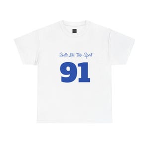 May include: A white short-sleeved t-shirt featuring the text Smells Like Teen Spirit in a blue script font above the large blue number 91 centered on the chest. The garment has a crew neckline and a small black label at the inner collar area.