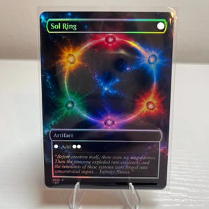 以下が含まれることがあります： Sol Ringというタイトルのトレーディングカード。宇宙をテーマにしたホログラフィックな背景に、6つの色付きオーブが配置された中央の光るリングが描かれています。カードにはArtifact、マナシンボル、インフィニティ・ストーンに関するフレーバーテキストが記載されています。