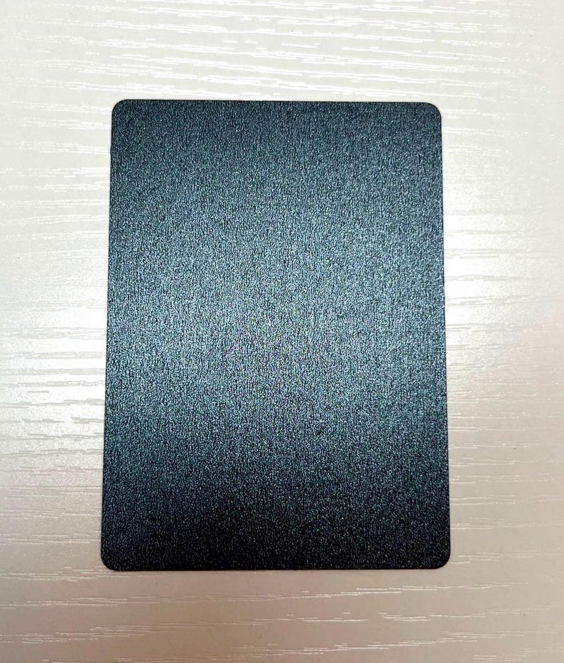 May include: A rectangular card with a dark blue, metallic, and textured finish. The surface has a fine, granular appearance that reflects light across its flat, uniform plane. The edges of the card are rounded, creating a smooth, consistent shape.