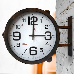 May include: A round wall-mounted clock with a dark brown metal frame and a white face featuring large black Arabic numerals. The clock has black hour and minute hands, with subtle rust-colored markings on the dial surface and a metal bracket.