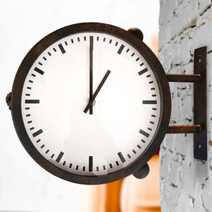 Pode incluir: Um relógio de parede redondo de face dupla com uma moldura de metal escuro e desgastado e um mostrador branco. O relógio apresenta marcas pretas simples para indicar horas e minutos, com dois ponteiros pretos marcando uma hora.