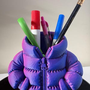 May include: A purple and blue gradient puffer jacket shaped pencil holder containing a green marker, a red marker, a pink pen, a blue marker, and a black pencil. The holder features a central zip detail and two side pockets, mimicking the design of a quilted winter coat.