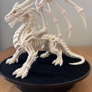 May include: An off-white, skeletal dragon figurine featuring a detailed bone structure, including a rib cage, spinal column, sharp claws, and large, extended wings. The creature is depicted in a standing position with its head turned slightly.