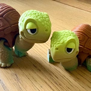 May include: Two turtle figurines with textured brown shells and light green heads and limbs. The larger turtle stands upright while the smaller turtle is positioned lower. Both figures feature detailed, sculpted skin patterns and large, sleepy eyes.