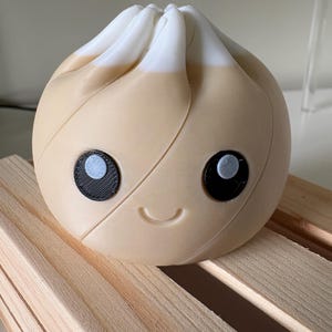 May include: A round, beige dumpling-shaped figure with a white top section. The object features two black circular eyes with white pupils and a small, curved, embossed mouth. The surface has a visible layered texture consistent with 3D printing.