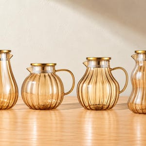 Puede incluir: Cuatro jarras de cristal de color ámbar con texturas acanaladas verticales y tapas de metal dorado. Cada jarra presenta un asa curva y una silueta distinta, que varía de formas redondeadas a alargadas, diseñadas para servir bebidas.