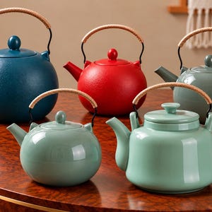 Puede incluir: Una colección de cinco teteras en varios colores, incluyendo azul oscuro, rojo, verde salvia y verde menta claro. Cada tetera presenta un cuerpo redondeado, un pico, una tapa con un pequeño pomo y un asa curva envuelta en material tostado.