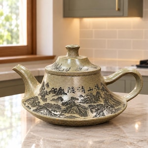 May include: A speckled light green ceramic teapot with a wide, low-profile base, a curved handle, and a matching lid. The body features a detailed black ink-style landscape illustration of mountains and buildings with Chinese calligraphy text.
