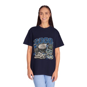 May include: A navy blue t-shirt featuring a graphic print with the text 2000s. The design includes illustrations of a flip phone, a portable CD player with headphones, a handheld gaming console, a pair of athletic sneakers, and a compact disc.