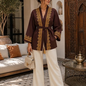 Peut inclure: Une veste ouverte marron foncé avec des motifs brodés dorés sur les revers et les poignets, accompagnée d'une ceinture à nouer assortie. L'ensemble comprend un pantalon large crème et un sac à main structuré crème avec une poignée.