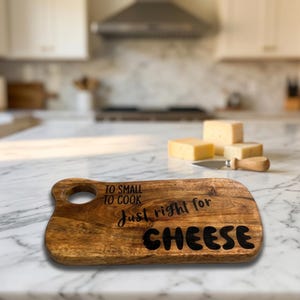 May include: A rectangular wooden serving board with a circular handle hole, featuring the black printed text: TO SMALL TO COOK, JUST RIGHT FOR CHEESE. The board has a natural brown wood grain finish and is positioned on a white marble surface.