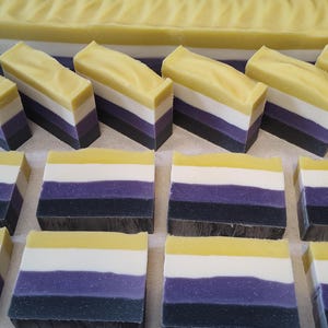 May include: Handmade soap bars in a non-binary pride flag design. The bars feature layers of yellow, white, purple, and black. A larger block of soap with a similar design is in the background. The soap is likely scented.