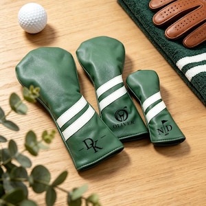Könnte beinhalten: Drei grüne Golfschlägerhauben aus Kunstleder, jeweils mit zwei weißen horizontalen Streifen versehen. Die Hauben sind mit schwarzem Text und Symbolen personalisiert, darunter die Initialen DK, der Name OLIVER und die Initialen ND.