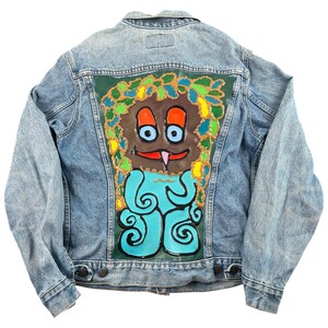 May include: Light blue denim jacket with a colorful, hand-painted design on the back. The artwork features a cartoonish creature with a brown face, blue eyes, and a red tongue, set against a green background. The creature has a blue body with swirling patterns.