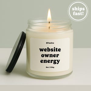 以下が含まれることがあります： 黒い蓋が付いた透明なガラス瓶に入った火のついたキャンドル。白いラベルには「8Twelve」「website owner energy」「8oz | 226g」という文字が印刷されています。キャンドルのワックスはオフホワイトで、明るい黄色の炎が温かい光を放っています。