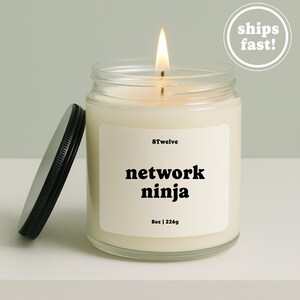 Peut inclure: Une bougie en pot de verre transparent avec une mèche allumée et un couvercle en métal noir posé à côté. L'étiquette blanche porte le texte 8Twelve, network ninja, et 8oz | 226g. La cire est blanc cassé et la flamme est jaune et orange.