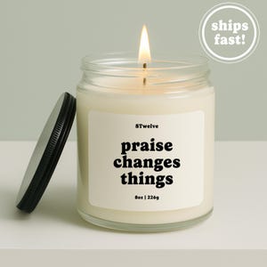 May include: A lit candle in a clear glass jar with a black metal lid. The white label features the text 8Twelve, praise changes things, and 8oz | 226g. The wax is off-white, and the flame is visible at the top of the wick in the center of the jar.