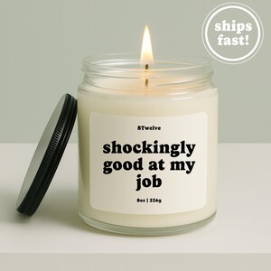May include: A clear glass candle jar with a lit wick and a black metal lid placed to the side. The white label features the text 8Twelve, shockingly good at my job, and 8oz | 226g. The wax is off-white and fills the jar to the top edge.