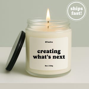 Op de afbeelding: Een kaars in een doorzichtige glazen pot met een brandende lont en een zwart metalen deksel ernaast. Het witte etiket bevat de tekst 8Twelve, creating what's next, en 8oz | 226g in zwarte letters. De gebroken witte was vult de cilindrische glazen houder.