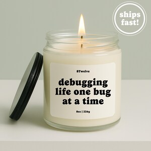 May include: A clear glass jar candle with a lit wick and a black metal lid placed to the side. The white label features the text 8Twelve, debugging life one bug at a time, and 8oz | 226g. The wax is off-white and fills the cylindrical glass container.