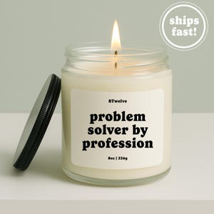 May include: A lit candle in a clear glass jar with a black lid. The white label features the text 8Twelve, problem solver by profession, and 8oz | 226g. The candle wax is off-white, and the flame is visible at the top of the glass container.