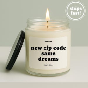 Könnte beinhalten: Eine brennende Kerze in einem klaren Glasgefäß mit schwarzem Deckel. Das Etikett trägt den Text 8Twelve, new zip code same dreams, und 8oz | 226g. Das Wachs ist cremeweiß und die Flamme leuchtet gelb. Die Kerze hat ein schlichtes, klares Design.