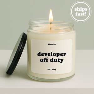 May include: A clear glass candle jar with a lit wick and a black metal lid resting beside it. The white label features the text 8Twelve, developer off duty, and 8oz | 226g. The wax is off-white, and the flame is bright yellow and orange.