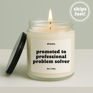 May include: A clear glass candle jar with a lit wick and a black metal lid resting beside it. The white label features the text 8Twelve, promoted to professional problem solver, and 8oz | 226g. The wax is off-white and fills the cylindrical container.