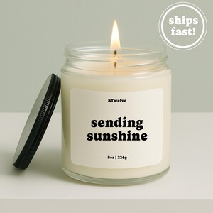 Op de afbeelding: Een brandende kaars in een doorzichtige glazen pot met een zwart metalen deksel ernaast. Het witte etiket bevat de tekst 8Twelve, sending sunshine, en 8oz | 226g. De was is gebroken wit en de vlam is zichtbaar aan de bovenkant van de lont.