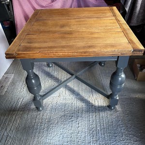 May include: A square table with a natural wood grain top and four dark grey painted legs. The legs feature a turned, bulbous design and are connected by a cross-shaped support structure underneath the tabletop surface.