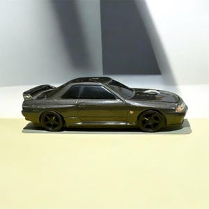 May include: A metallic dark gray toy car featuring a two-door coupe body style, a rear spoiler, black tinted windows, and five-spoke black wheels. The vehicle has a low-profile design with a small emblem on the front fender and a sleek hood.