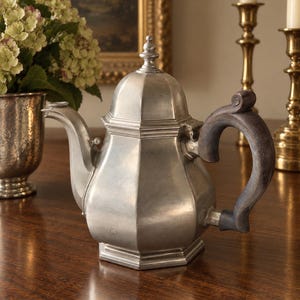 May include: A metallic teapot with a hexagonal body, a curved spout, and a dark, textured wooden handle. The teapot features a matching lid with a small finial on top. The surface of the metal has a matte, brushed finish with visible seams.