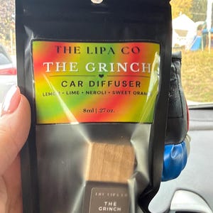 May include: A black pouch containing a car diffuser with a label reading THE LIPA CO THE GRINCH CAR DIFFUSER LEMON LIME NEROLI SWEET ORANGE 8ml .27 oz. The diffuser has a wooden cap and a small glass bottle with a label reading THE GRINCH CAR DIFFUSER 8 mL.