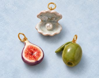 Puede incluir: Tres pequeños dijes con anillas doradas: una concha de cerámica abierta que contiene una perla blanca, un higo cortado por la mitad que muestra pulpa roja y blanca con piel morada, y una aceituna verde brillante con una pequeña hoja.