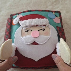 Pode incluir: Um livro de feltro feito à mão com um rosto de Papai Noel na capa. O Papai Noel tem um gorro vermelho com acabamento branco, barba branca, nariz rosa e olhos fechados. O fundo é azul-petróleo com pequenos enfeites de feltro e borda xadrez.